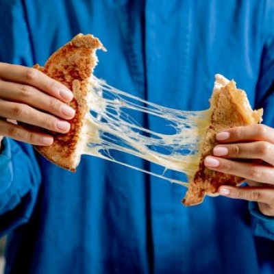 A person in a blue button-down shirt is pulling apart a toasted cheese sandwich, with strings of melted cheese between the two sides.
